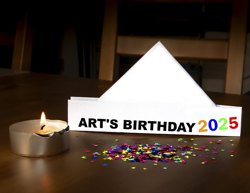 Art's Birthday - Geburtstag der Kunst – Robert Filliou und der Anniversaire de l’art. Kuriose Feiertage - 17. Januar © 2025 Sven Giese Tisch mit brennendem Teelicht, buntem Konfetti und Papierhut mit der Aufschrift "Art's Birthday 2025".