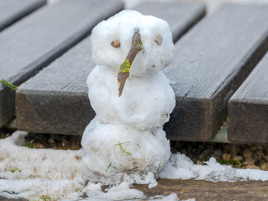 Kleiner Schneemann aus drei Schneekugeln mit Zweig-Nase steht vor grauer Holzbank im Freien.