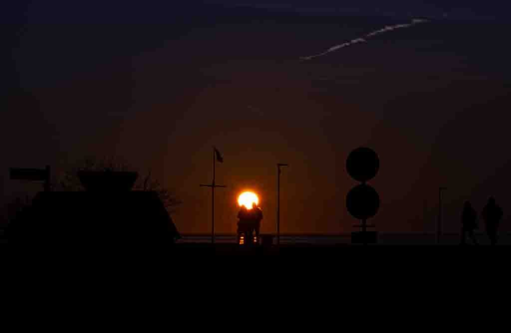 Zwei Personen stehen Arm in Arm vor untergehender Sonne, dunkle Silhouetten auf abendlicher Promenade.