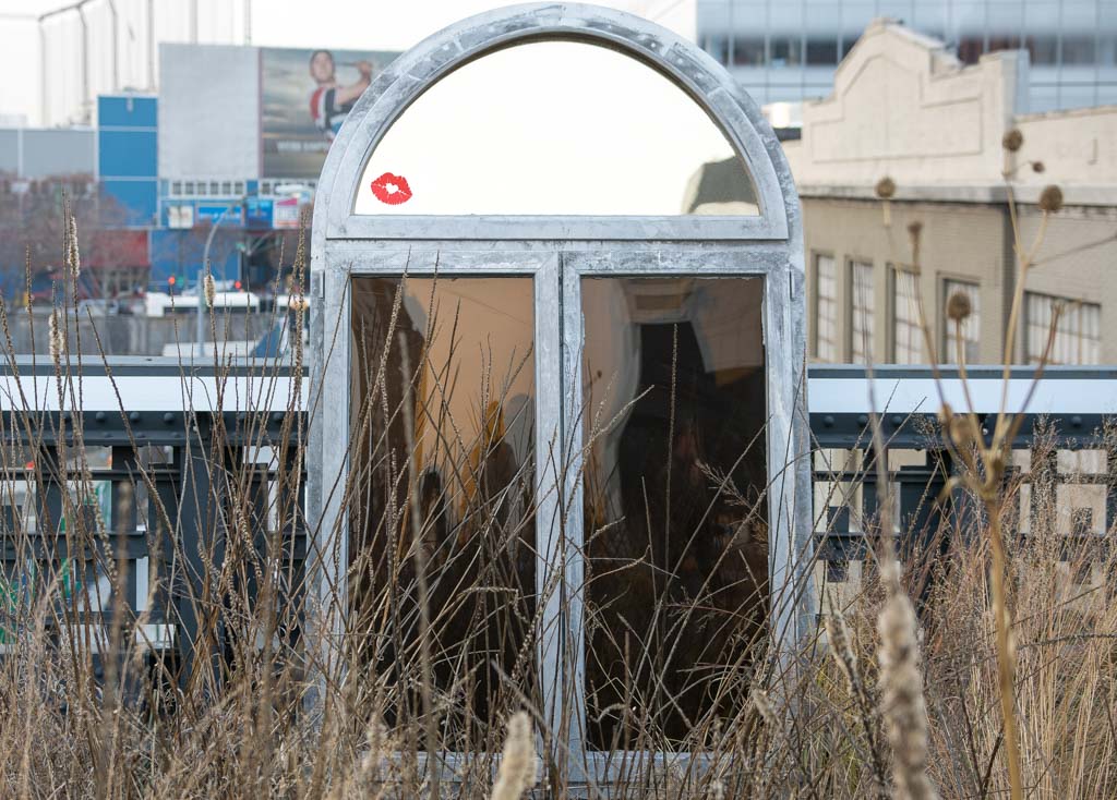 Spiegel mit Kussmund-Aufkleber steht in einem wild bewachsenen Außenbereich.