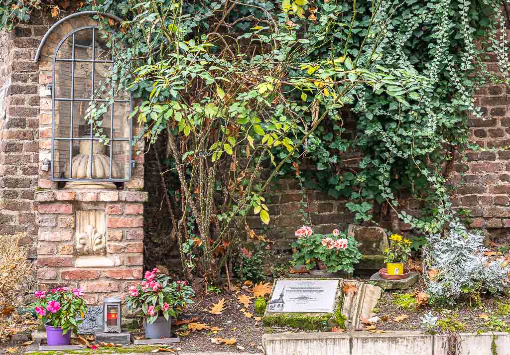 Mauer mit Bildstock der heiligen Getrud von Nivelles, Blumen, Kerzen und Gedenktafel im Vordergrund.