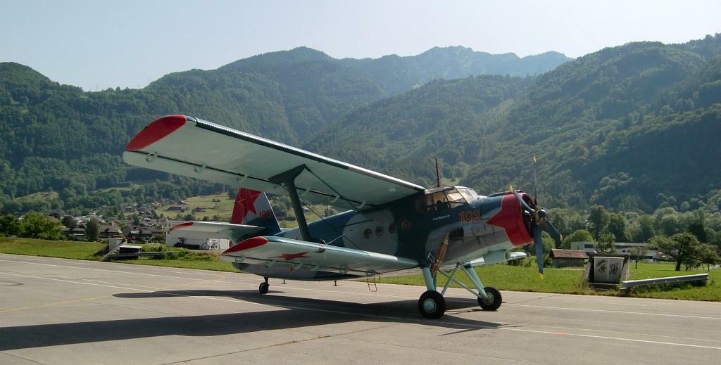Blau-silber-rotes Doppeldecker-Oldtimer-Flugzeug auf Rollfeld mit Bergen und Wiesen im Hintergrund.