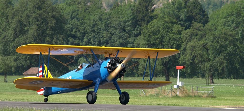 Blau-gelbes Doppeldecker-Oldtimer-Flugzeug auf Rollfeld mit Bergen und Wiesen im Hintergrund.