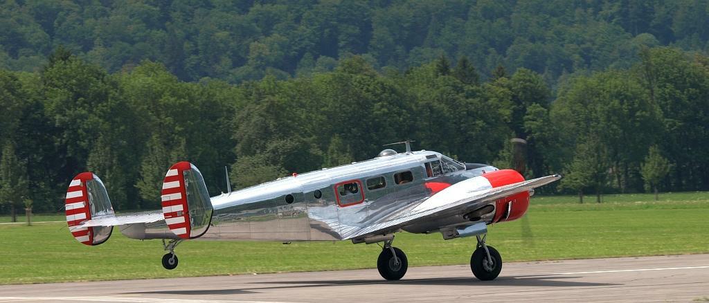 Retro-Zweimotor-Flugzeug in Silber mit rot-weißem Leitwerk landet auf einer Wiese vor Waldkulisse.
