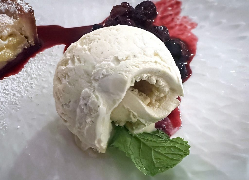 National Vanilla Ice Cream Day – Vanilleeis-Tag in den USA. Kuriose Feiertage – 23. Juli © 2026 Sven Giese – Bild 1 Kugel Vanilleeis mit Beerensauce und Minze auf weißem Teller, leicht angeschmolzen.