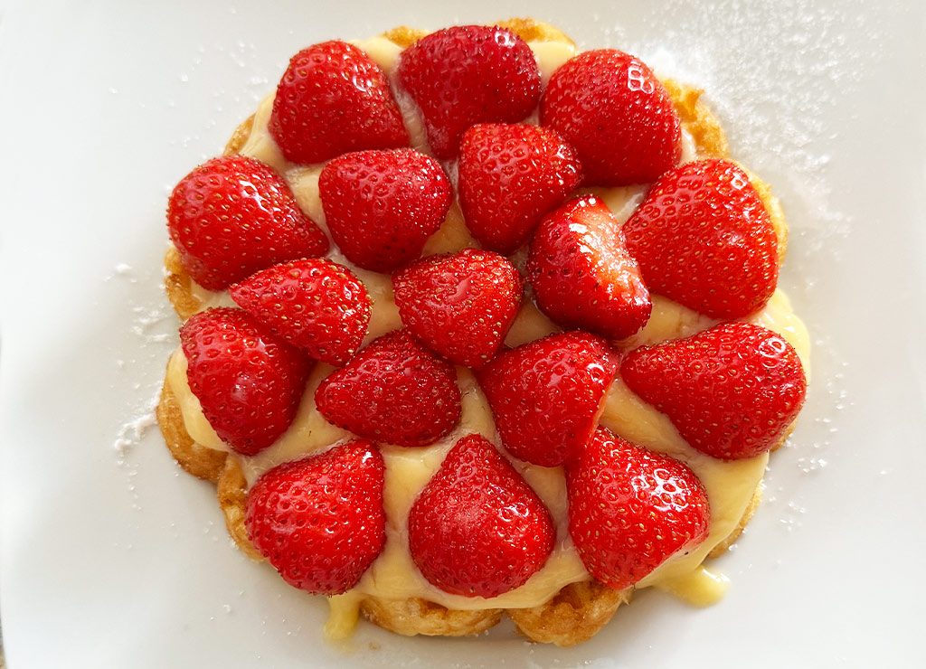 Eine belgische Waffel, bedeckt mit Vanillecreme und Erdbeeren, auf einem weißen Teller serviert.