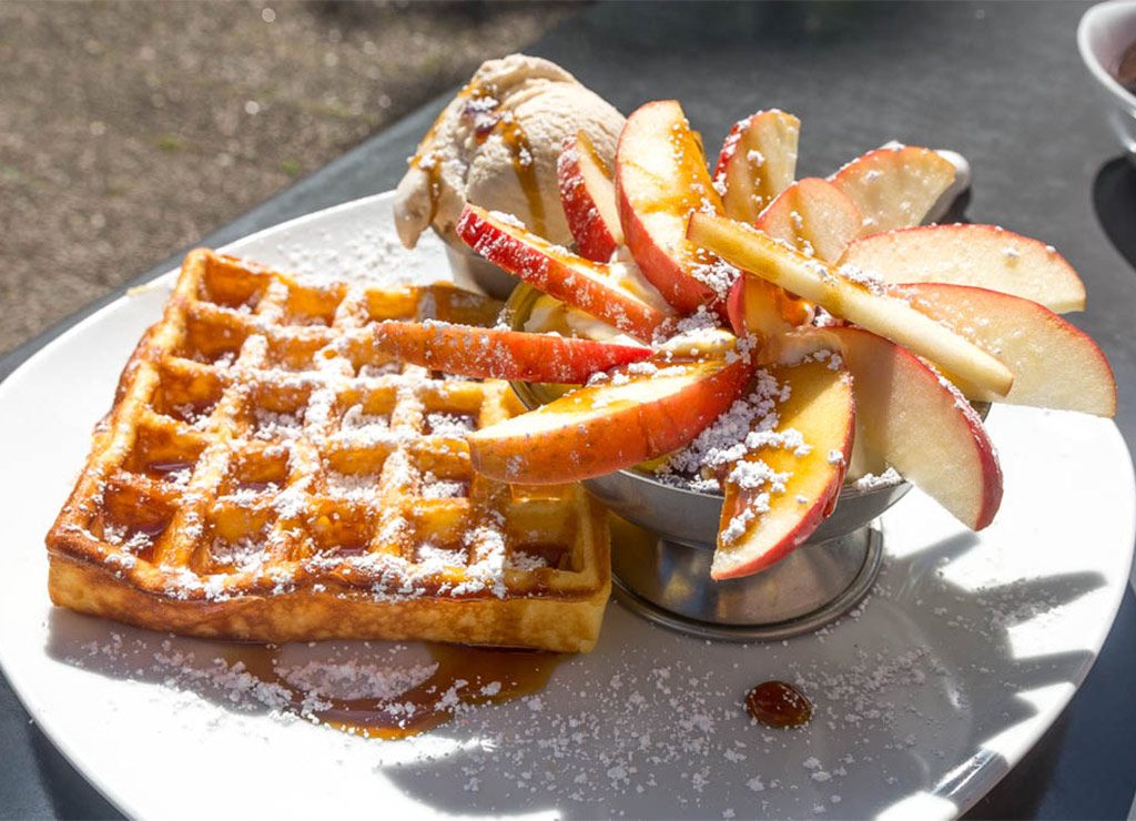 National Waffle Day - Tag der Waffel in den USA. Kuriose Feiertage - 24. August © 2022 Sven Giese - Bild 1 Eine frische Waffel mit Apfelscheiben, Puderzucker und einer Kugel Eis, serviert auf einem weißen Teller.