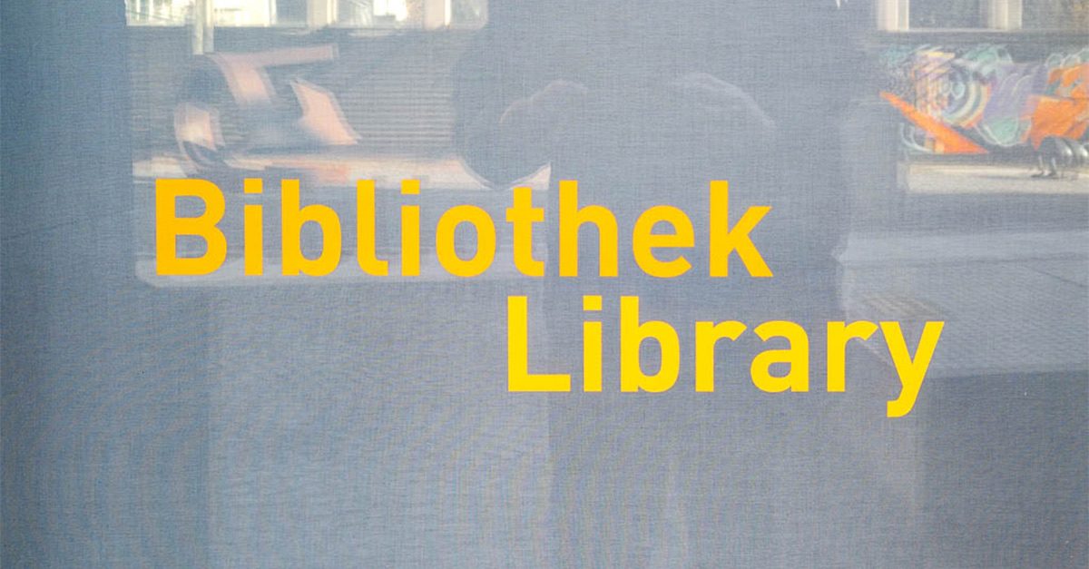 Fenster mit gelber Aufschrift "Bibliothek Library", Spiegelung von Gebäuden im Hintergrund.