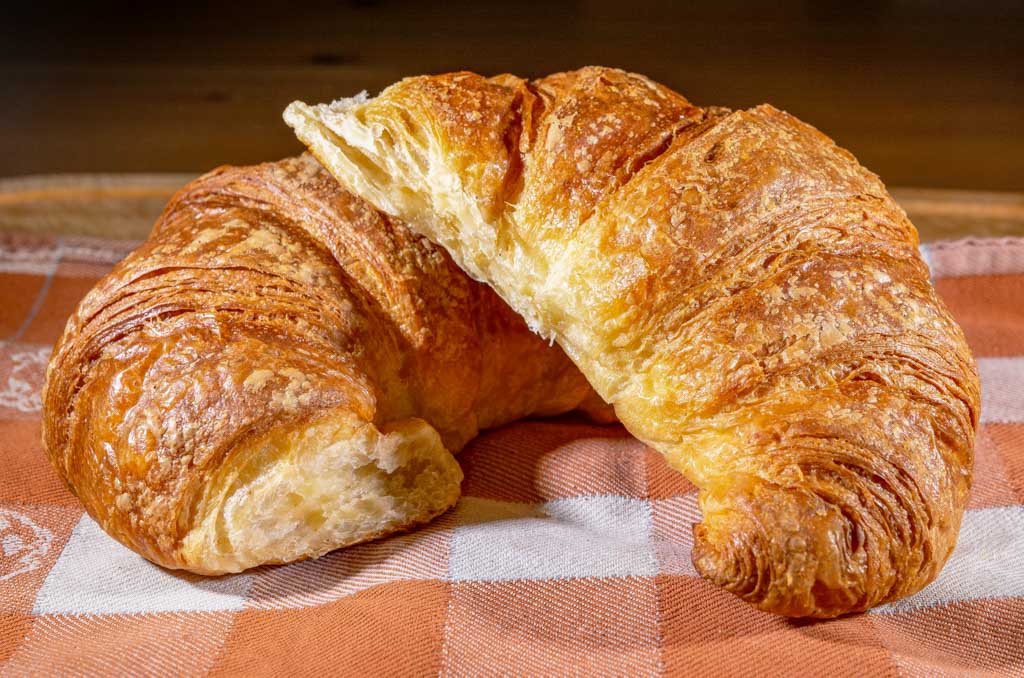 National Croissant Day - Tag des Croissants in den USA. Kuriose Feiertage - 30. Januar © 2022 Sven Giese Zwei frische Croissants liegen gestapelt auf einer karierten Tischdecke eines Holztisches.