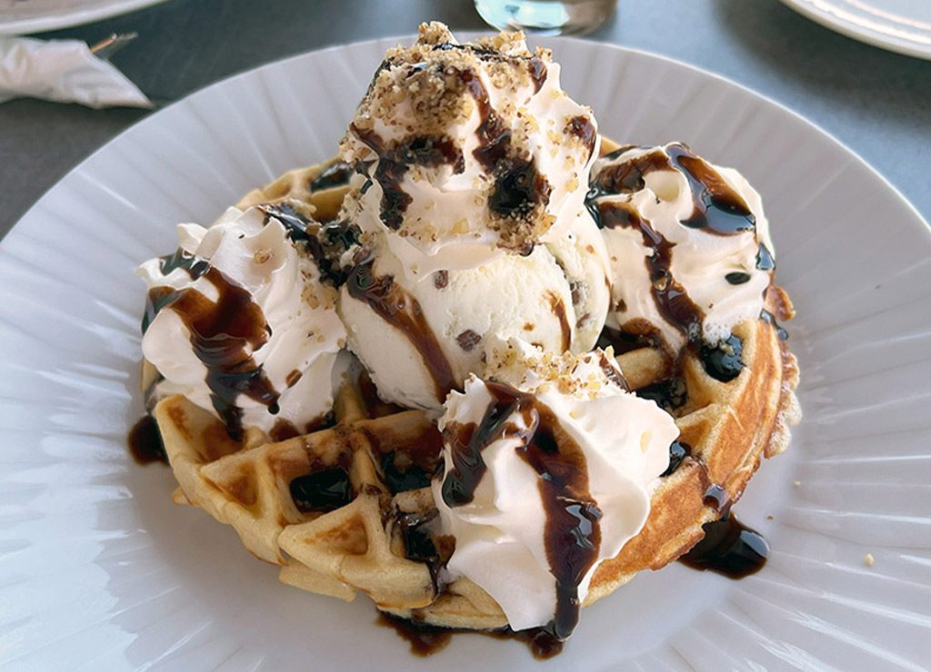 Våffeldagen – Tag der Waffel in Schweden. Kuriose Feiertage - 25. März © 2024 Sven Giese. Waffel mit Vanilleeis, Sahne, Schokosauce und Nüssen auf weißem Teller kunstvoll serviert.