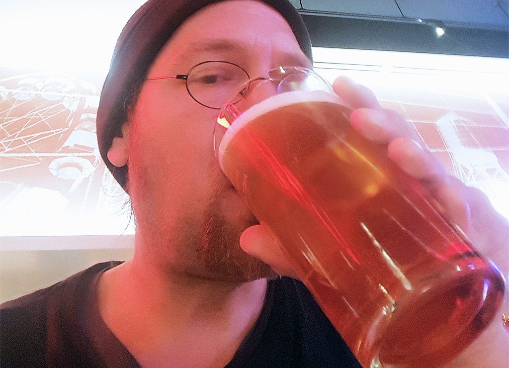 Mann mit Mütze und Brille trinkt Bier aus Glas in Innenraum mit roter Beleuchtung.