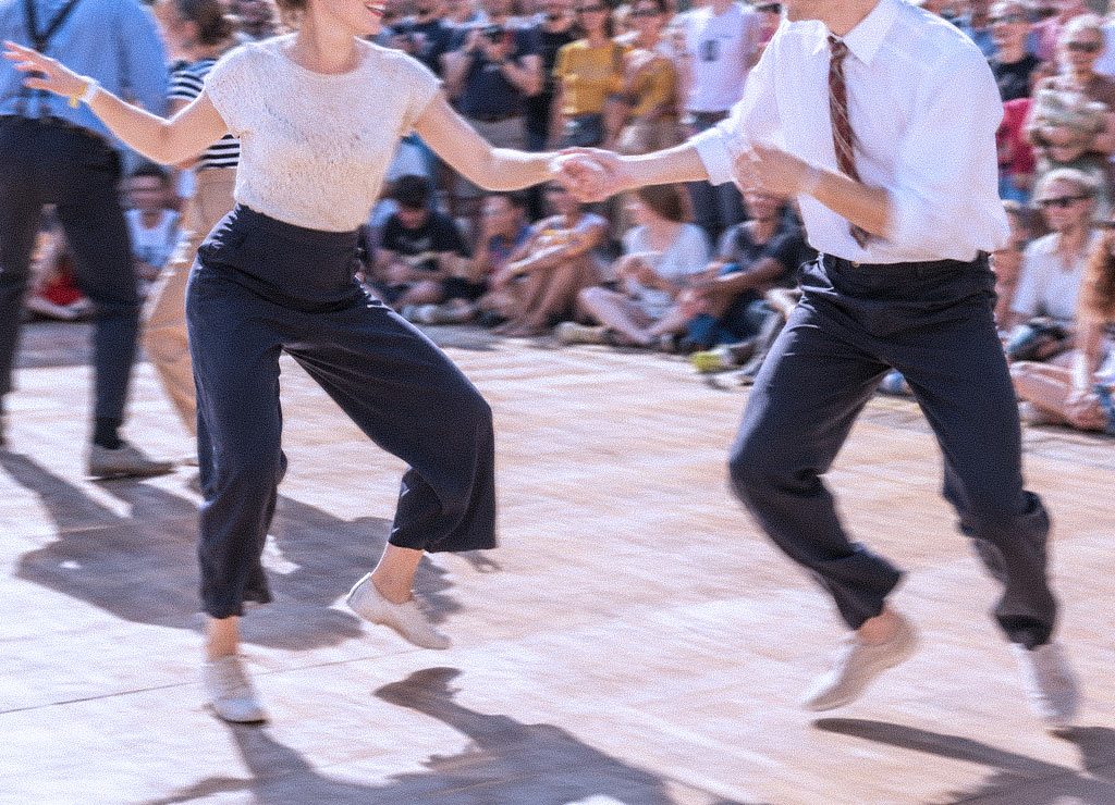 Lindy-Hop-Paar springt im hellen Tageslicht, Zuschauer sitzen unscharf im Hintergrund.