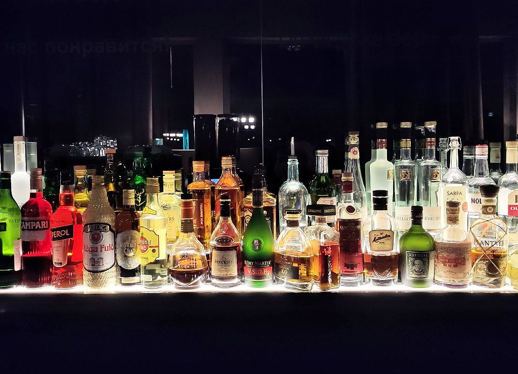 Name Your Poison Day – der US-amerikanische Was-willst-Du-trinken-Tag? Kuriose Feiertage - 8. Juni © 2025 Sven Giese Vielzahl beleuchteter Spirituosenflaschen auf einer Bar, darunter Rum, Whiskey und Likör.