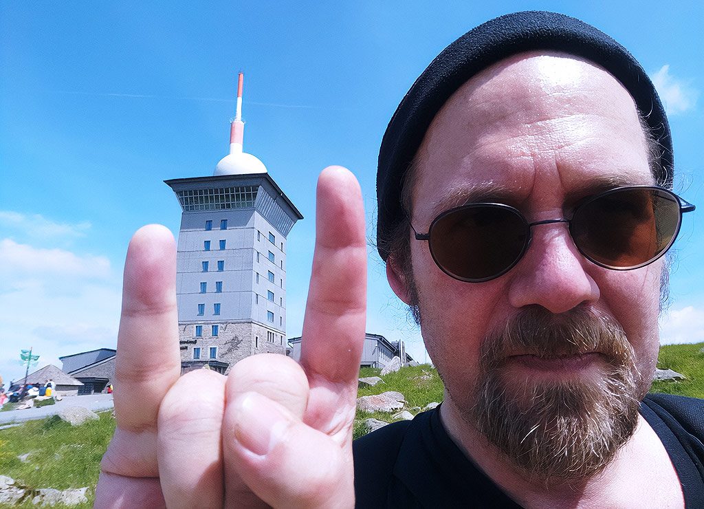 National Sun Glasses Day – Tag der Sonnenbrille in den USA. Kuriose Feiertage - 27. Juni © 2022 Sven Giese Mann mit Bart, Mütze und Sonnenbrille zeigt Rock-Geste vor Turm im Hintergrund.