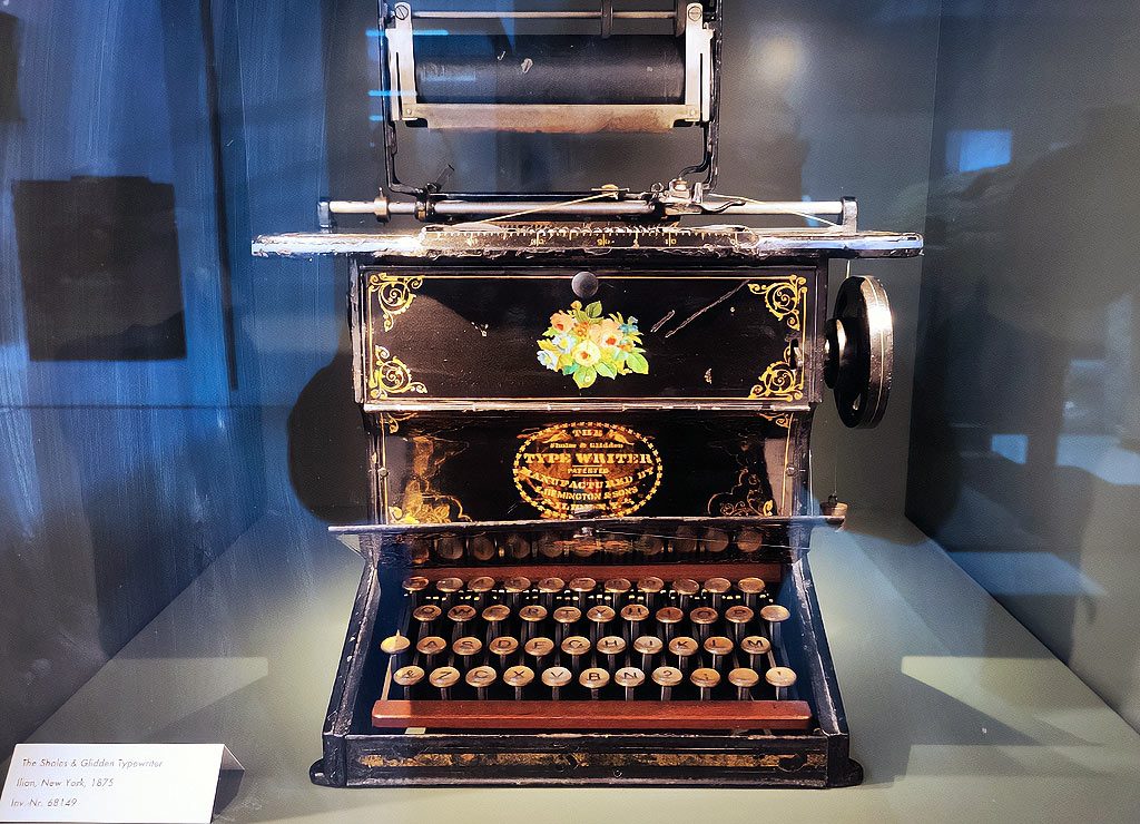 Typewriter Day - internationaler Tag der Schreibmaschine. Kuriose Feiertage - 23. Juni © 2026 Sven Giese - Bild 4 Sholes & Glidden Typewriter mit Blumenmotiv und Rundtasten steht hinter Glas im Deutschen Museum, München.