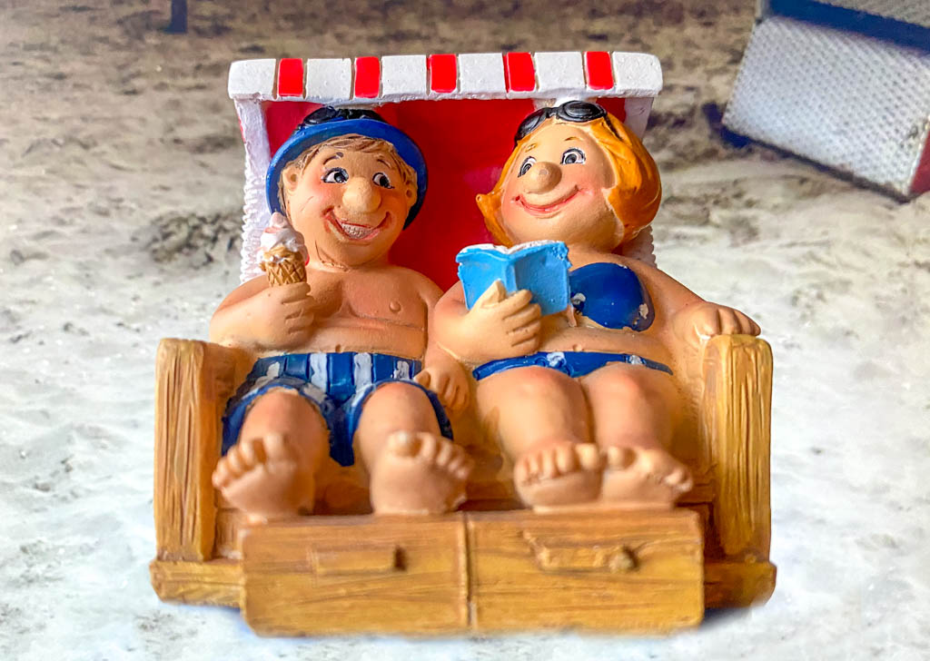 National Bikini Day - Tag des Bikinis in den USA. Kuriose Feiertage - 5. Juli © 2021 Dietmar Giese Keramikfiguren eines Pärchens im Strandkorb, er in Badehose und mit Eis, sie im Bikini und mit Buch in der Hand.