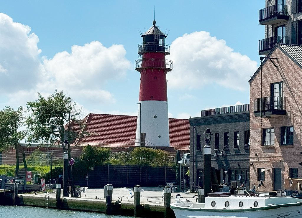 Ein rot-weißer Leuchtturm steht zwischen modernen Gebäuden am Ufer eines Flusses, mit einem Boot im Vordergrund.