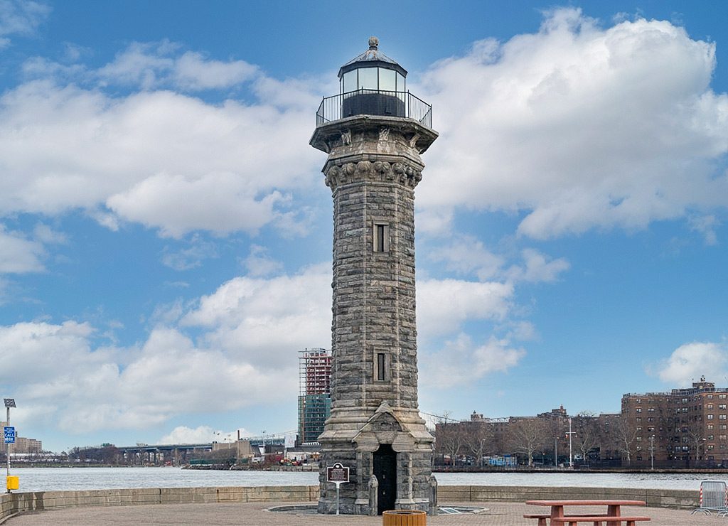 National Lighthouse Day – Tag des Leuchtturms in den USA. Kuriose Feiertage - 7. August © 2024 Sven Giese – Bild 1 Ein historischer Steinturm-Leuchtturm steht auf einer gepflasterten Fläche mit einem Fluss und Stadtgebäuden im Hintergrund.Konkret handelt es sich um den Leuchtturm Blackwell Island Light auf Roosevelt Island in New York.