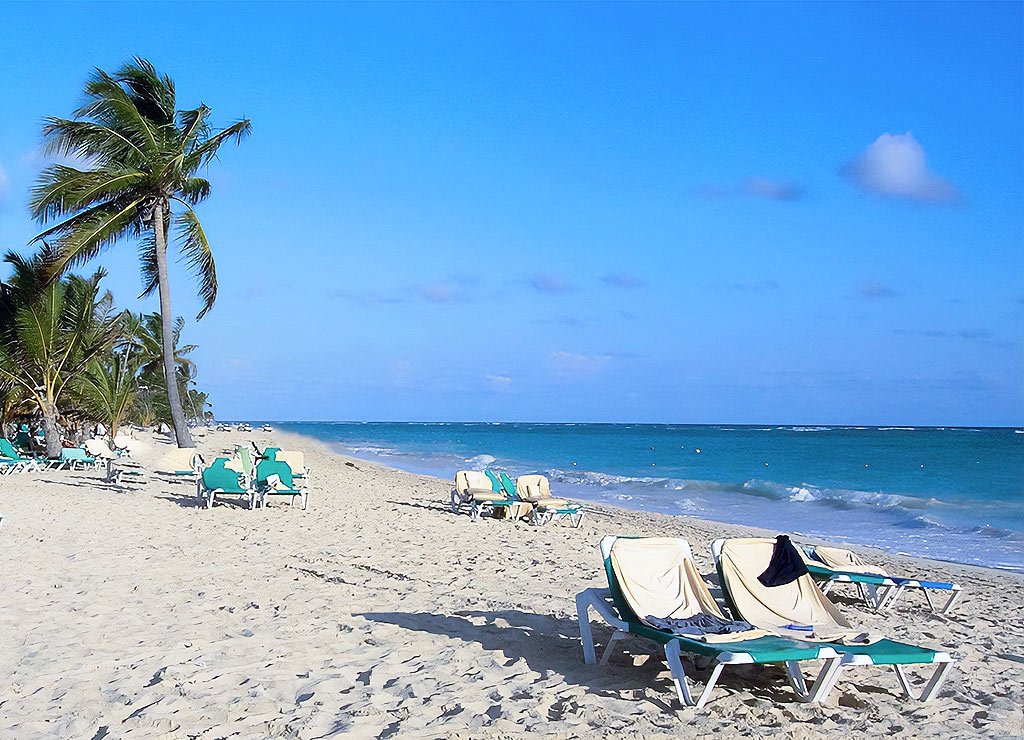 National Relaxation Day - Tag der Erholung in den USA. Kuriose Feiertage - 15. August © 2021 Sven Giese - Bild 1 Ein ruhiger Sandstrand mit leeren Liegestühlen und Palmen, die sich im Wind wiegen. Der blaue Himmel und das türkisfarbene Meer erstrecken sich in die Ferne.