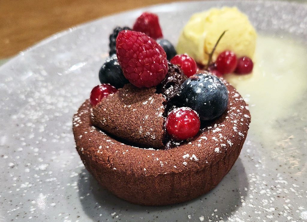 Schokoladenküchlein mit flüssigem Kern, Beeren und Puderzucker, Vanilleeis im Hintergrund, serviert auf grauem Teller.