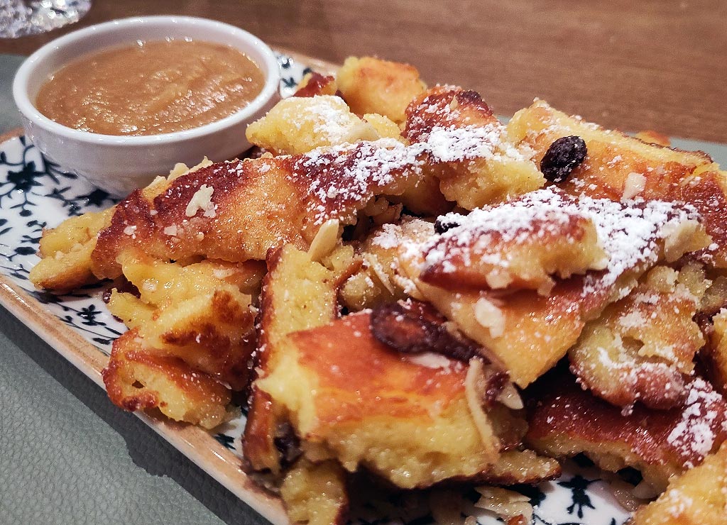 Frischer Kaiserschmarrn mit Rosinen und Mandeln, bestäubt mit Puderzucker, dazu Apfelmus im Schälchen.
