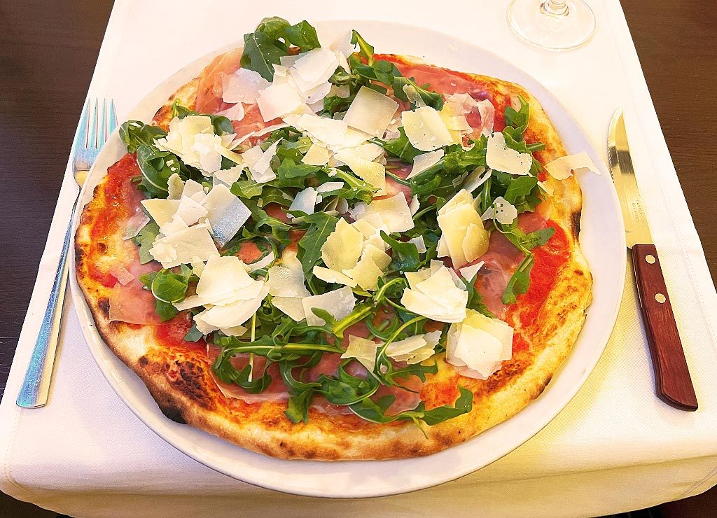 Pizza belegt mit frischem Rucola, Schinken und Parmesanflocken, serviert auf gedecktem Tisch.
