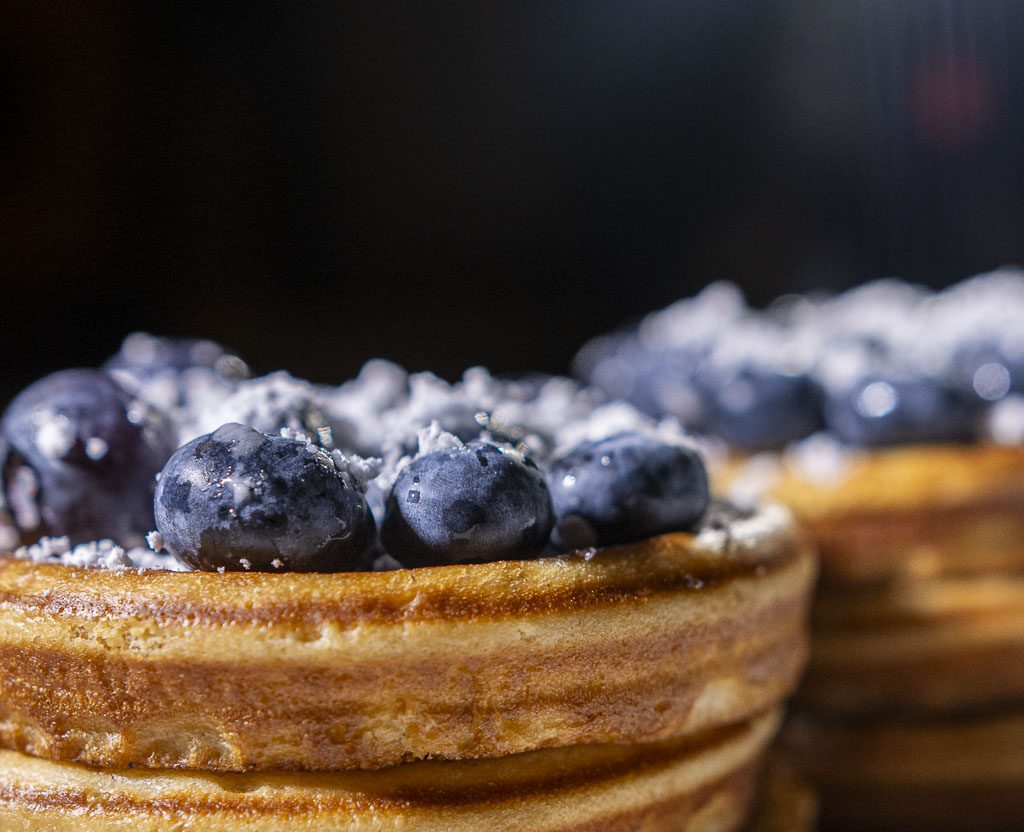 National Blueberry Pancake Day – Tag des Blaubeerpfannkuchens in den USA. Kuriose Feiertage - 28. Januar © 2025 Sven Giese – Bild 3 Nahaufnahme von gestapelten Pfannkuchen mit frischen Blaubeeren und Puderzucker vor dunklem Hintergrund.