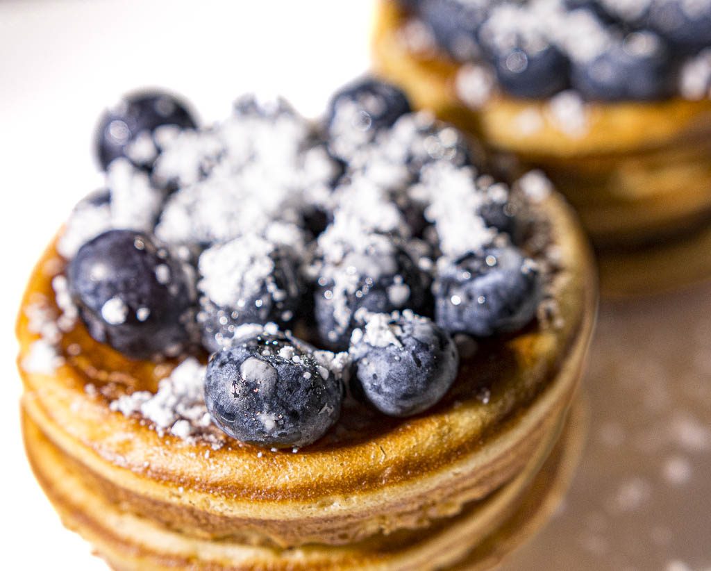 National Blueberry Pancake Day – Tag des Blaubeerpfannkuchens in den USA. Kuriose Feiertage - 28. Januar © 2025 Sven Giese – Bild 1 Zwei gestapelte Pfannkuchen mit frischen Blaubeeren und Puderzucker bestreut, Nahaufnahme.