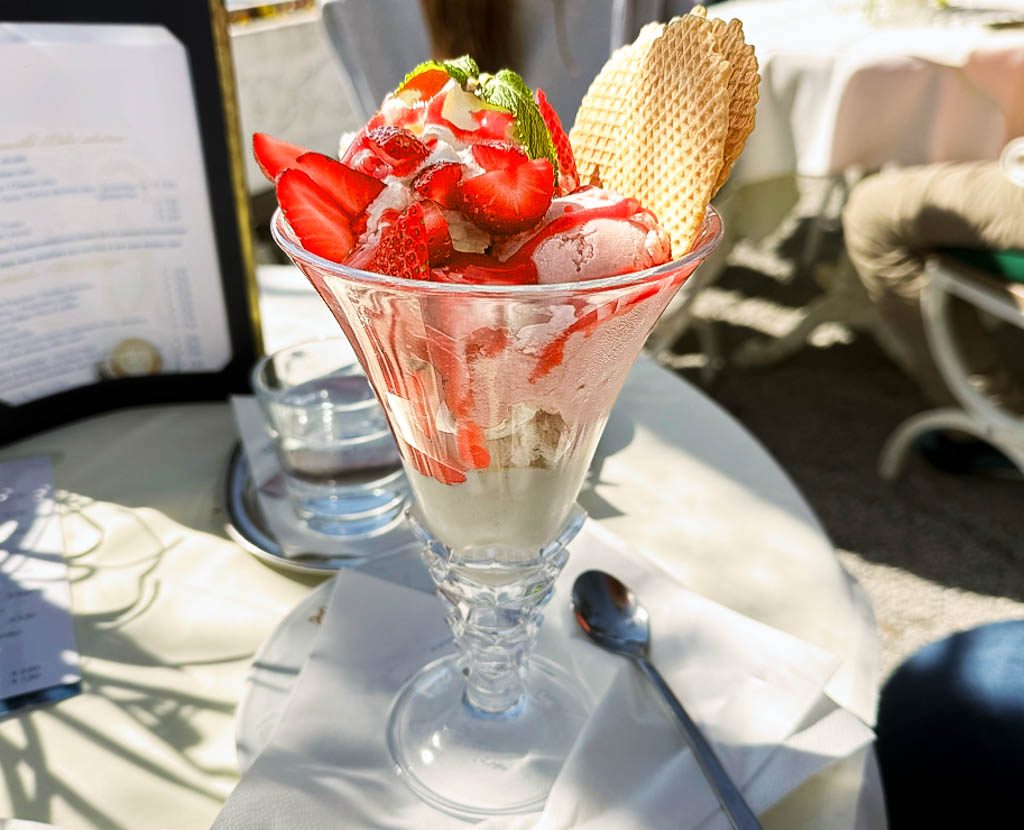 National Strawberry Ice Cream Day - Erdbeereis-Tag in den USA. Kuriose Feiertage - 15. Januar © 2024 Sven Giese "Ein Eisbecher mit Erdbeeren, Sahne, Minze und Waffeln auf einem gedeckten Tisch im Freien.