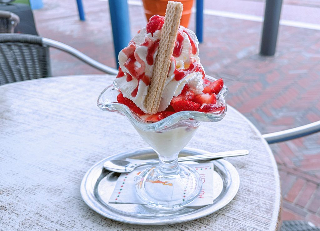 Eisbecher mit frischen Erdbeeren, Schlagsahne, roter Erdbeersauce und Waffel auf einem Cafétisch.