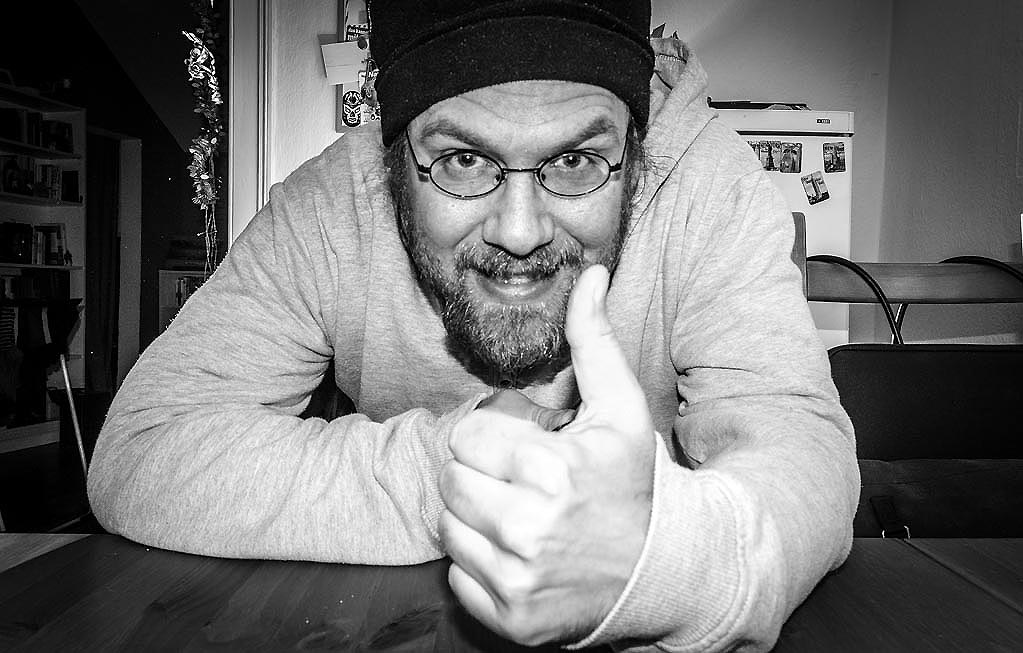 Everything You Do Is Right Day - der amerikanische Alles-was-Du-machst-ist-richtig-Tag. Kuriose Feiertage - 16. März © 2020 Sven Giese Schwarzweißbild einer lächelnden Person mit Daumen-hoch-Geste am Tisch in Innenraum.