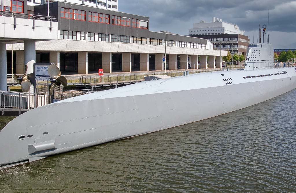 Großes weißes U-Boot liegt vertäut im Wasser, im Hintergrund moderne Gebäude und Propeller.