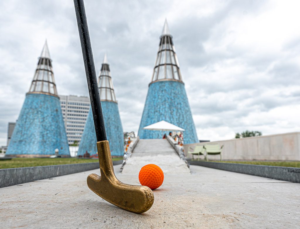 National Miniature Golf Day – Nationaler Minigolf-Tag in den USA. Kuriose Feiertage – zweiter Samstag im Mai © 2026 Sven Giese - Bild 1 Minigolfschläger und oranger Ball auf Bahn, dahinter ragen drei blaue Hindernistürme.