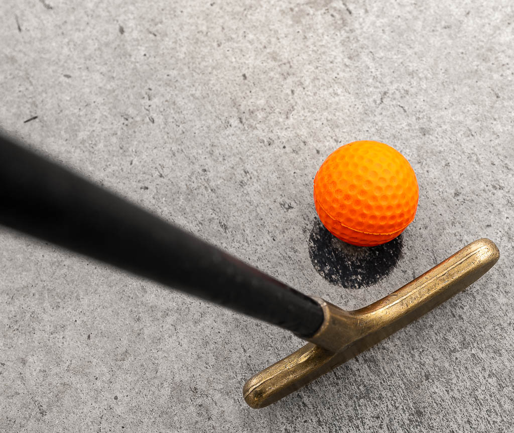 National Miniature Golf Day – Nationaler Minigolf-Tag in den USA. Kuriose Feiertage – zweiter Samstag im Mai © 2026 Sven Giese - Bild 3 Goldener Minigolfschläger liegt neben orangem Ball auf grauer Betonbahn von oben.