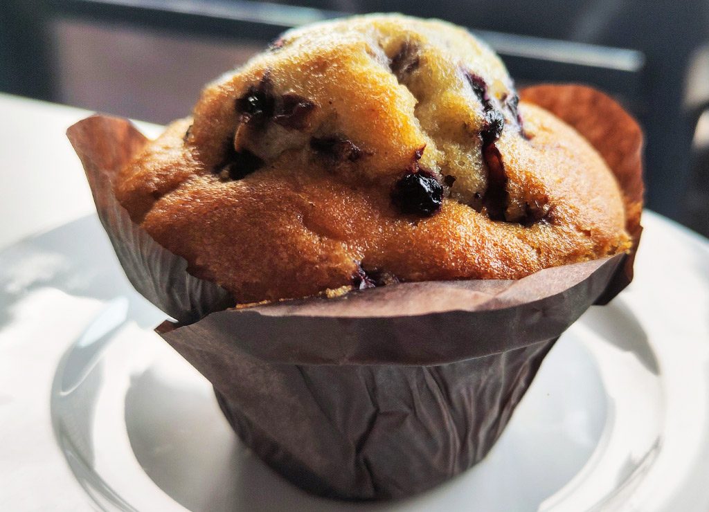 National Blueberry Muffin Day – Tag des Blaubeermuffins in den USA. Kuriose Feiertage – 11. Juli © 2026 Sven Giese Goldbrauner Blaubeermuffin mit sichtbaren Beeren steht in brauner Papierform auf weißem Teller.