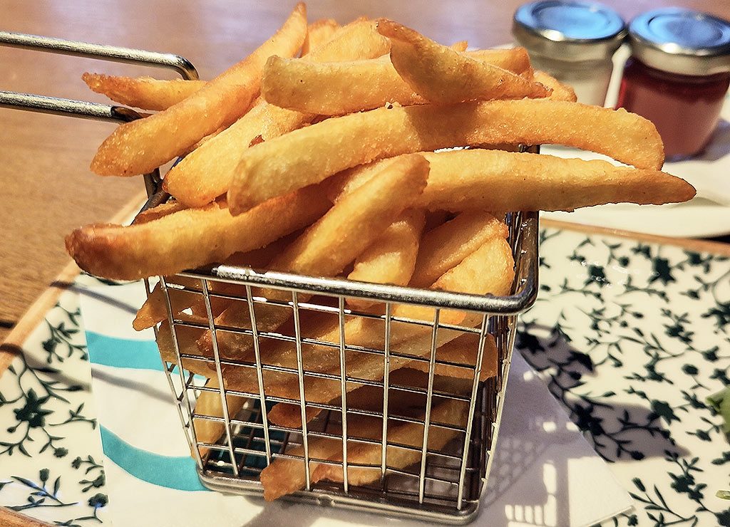 National French Fries Day – Pommes-Tag in den USA. Kuriose Feiertage – 13. Juli © 2026 Sven Giese – Bild 1 Goldbraune Pommes frites liegen in kleinem Metallkorb auf gemustertem Teller neben zwei Dips.