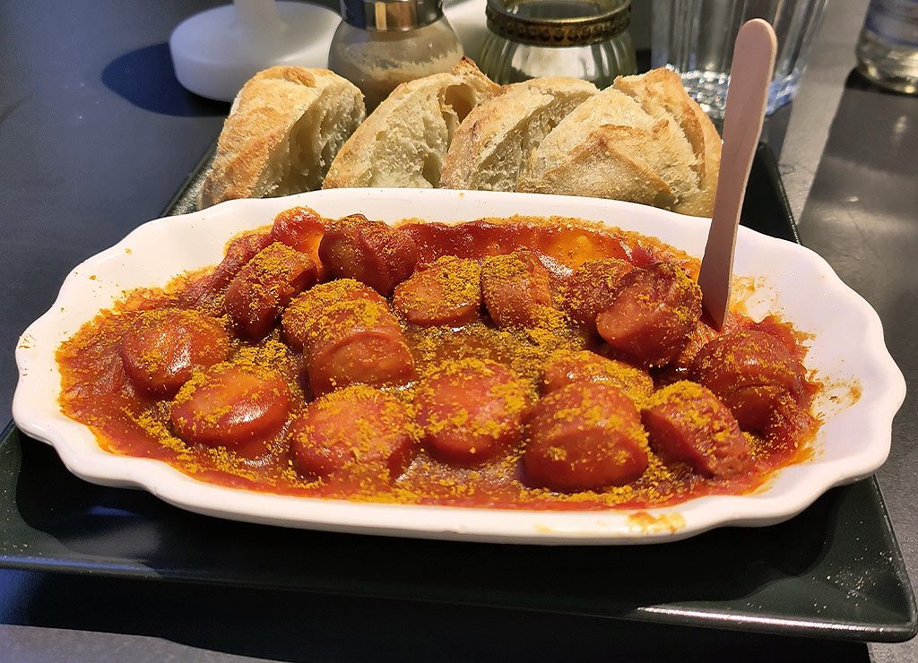 Tag der Currywurst. Kuriose Feiertage – 4. September © 2026 Sven Giese – Bild 4 Currywurststücke in roter Sauce mit Currypulver, dazu knusprige Baguettescheiben auf schwarzem Tablett.