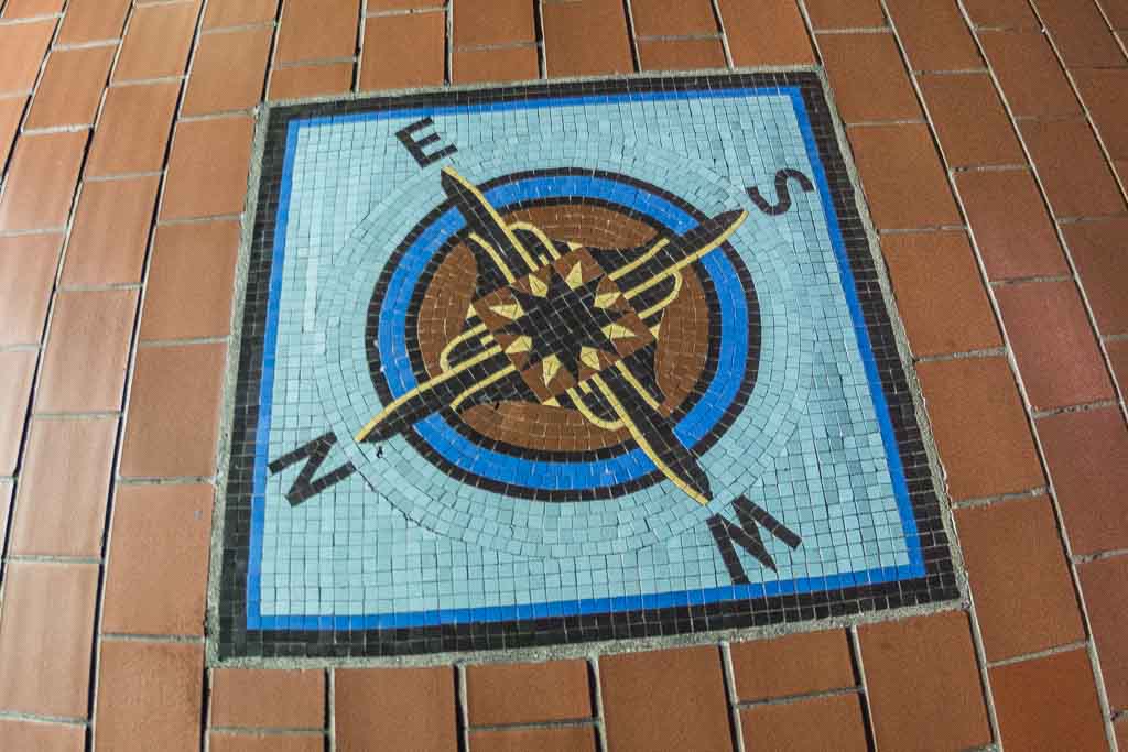 Ein Kompass-Mosaik mit Himmelsrichtungen auf einem terrakottafarbenen Boden.