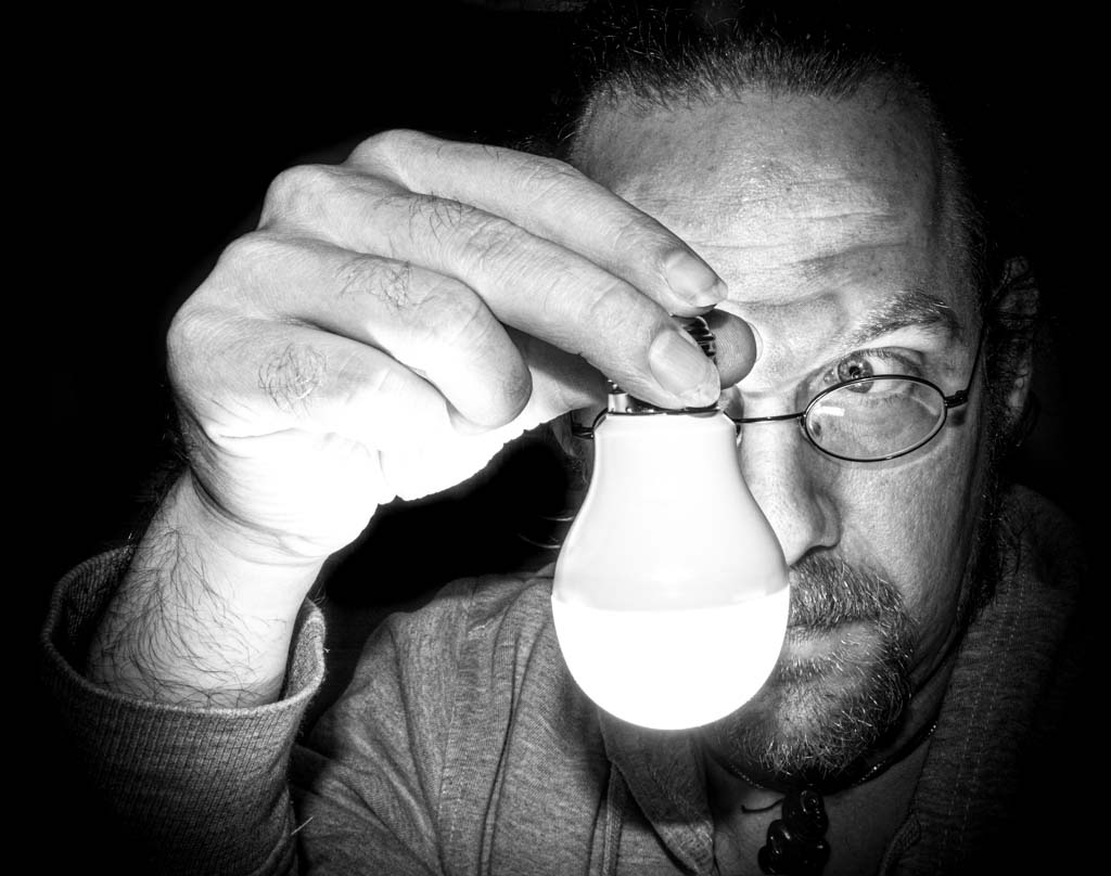 Be Electrific Day - Sei-elektrisierend-Tag in den USA. Kuriose Feiertage - 11. Februar © 2021 Sven Giese Ein Mann hält eine leuchtende Glühbirne vor sein Gesicht, das teils im Schatten liegt.