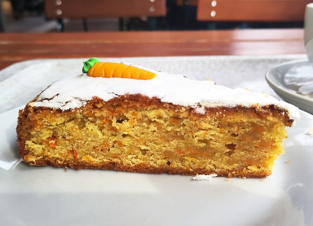 National Carrot Cake Day – Tag des Karottenkuchens in den USA. Kuriose Feiertage 3. Februar © 2025 Sven Giese – Bild 2 Ein Stück Karottenkuchen mit weißem Zuckerguss und einer kleinen Marzipan-Möhre auf einem Teller.