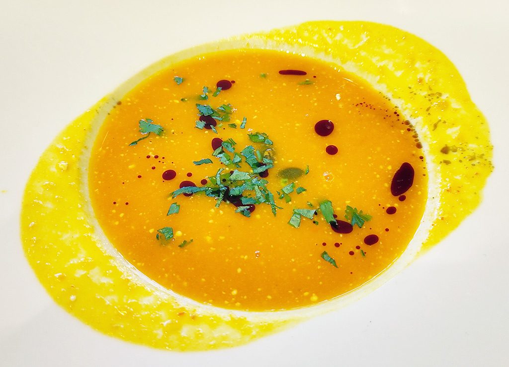 Cremige Kürbissuppe mit Kräutern und roten Öltropfen, serviert auf einem weißen Teller mit gelbem Rand
