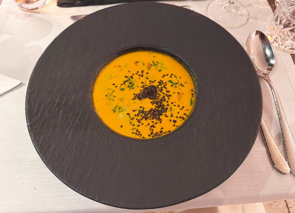 Dunkler Teller mit cremiger Kürbissuppe, garniert mit schwarzen Sesamsamen und Kräutern auf weißer Tischdecke.