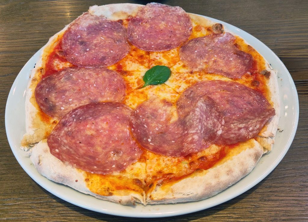 National Pizza Day – Tag der Pizza in den USA. Kuriose Feiertage – 9. Februar © 2026 Sven Giese – Bild 3 National-Pizza-Tag.jpg: Runde Salami-Pizza auf weißem Teller, knuspriger Rand, Basilikumblatt als Deko in der Mitte.
