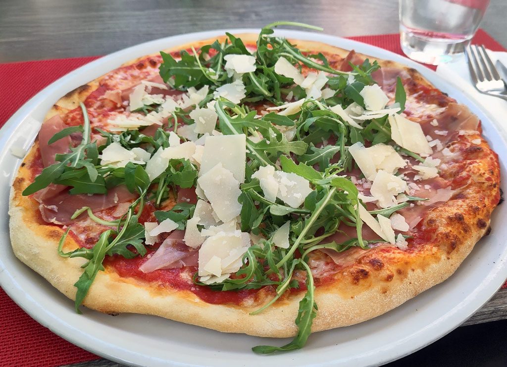 National Pizza Day – Tag der Pizza in den USA. Kuriose Feiertage – 9. Februar © 2026 Sven Giese – Bild 4 Pizza mit Rucola, Prosciutto und Parmesanspänen auf Teller, Glas und Besteck daneben.