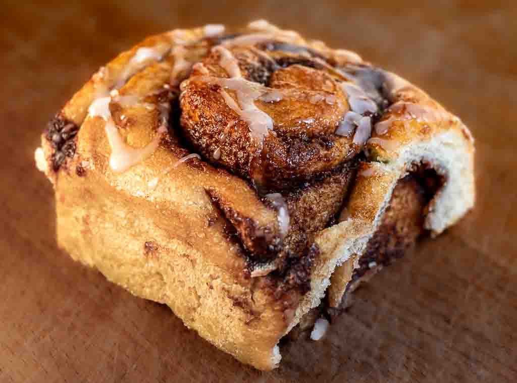 National Sticky Bun Day – Tag der süßen Schnecken in den USA. Kuriose Feiertage - 21. Februar © 2025 Sven Giese – Bild 1 Angeschnittene Zimtschnecke mit klebriger Zimtfüllung und süßer Glasur auf Holzuntergrund.