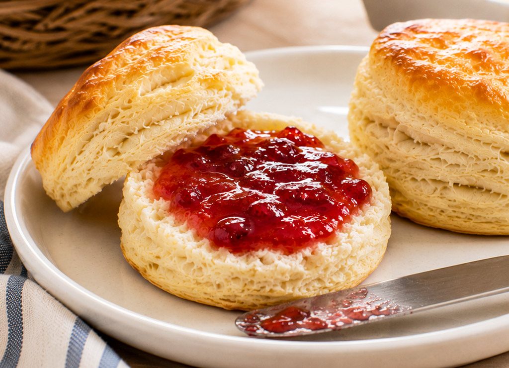 Aufgeschnittener Buttermilk Biscuit mit roter Marmelade und Messer auf hellem Frühstücksteller.