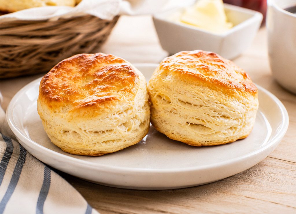 Zwei goldbraune Buttermilk Biscuits auf weißem Teller vor hellem Frühstückshintergrund.