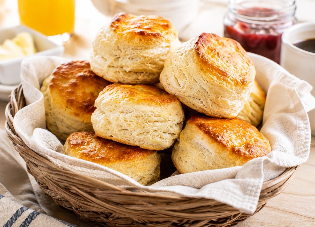 Korb mit goldbraunen Buttermilk Biscuits auf hellem Frühstückstisch mit Butter, Marmelade und Kaffee.