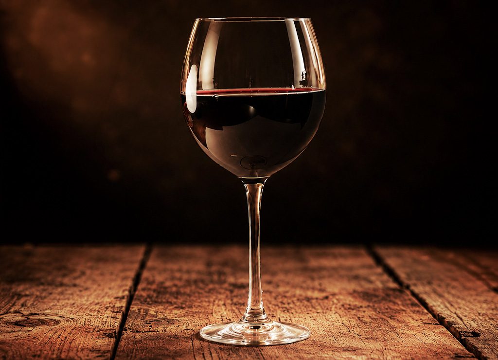 National Wine Day – Tag des Weins in den USA. Kuriose Feiertage – 25. Mai © 2026 Sven Giese - Bild 1 Rotweinglas steht auf rustikalem Holztisch vor dunklem Hintergrund im warmen Licht.