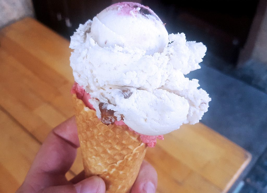 National Ice Cream Day – Tag des Eiscreme in den USA. Kuriose Feiertage – dritter Sonntag im Juli (c) 2025 Sven Giese – Bild 3 Hand hält Eiswaffel mit zwei Kugeln Eis, Vanille mit Stückchen und darunter rosa Fruchtsorte.